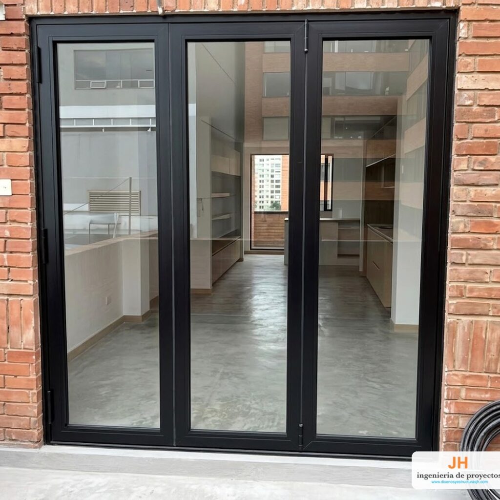 Puerta plegable europea AWA (Eurovitral) de tres hojas en aluminio negro y vidrio templado, salida a terraza de apartamento en Bogotá, instalada por JH