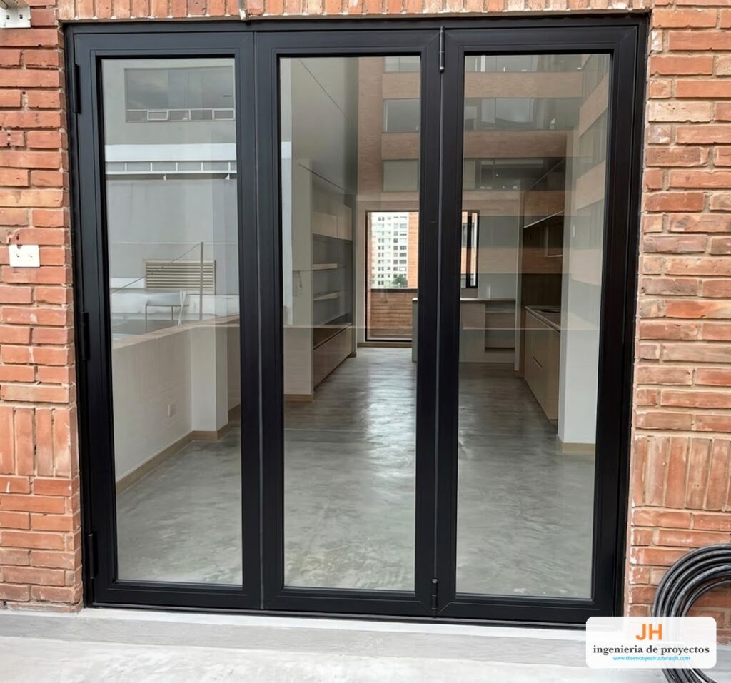 Puerta plegable europea AWA (Eurovitral) de tres hojas en aluminio negro y vidrio templado, salida a terraza de apartamento en Bogotá, instalada por JH