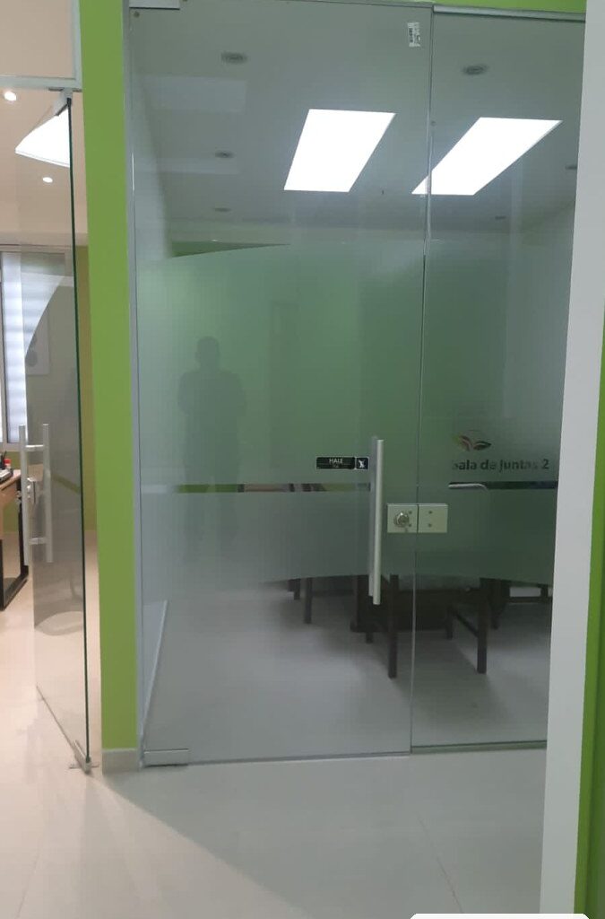 Puerta batiente en vidrio templado con franja esmerilada, manija tubular y chapa en acero inoxidable, sala de juntas en Bogotá, obra de JH